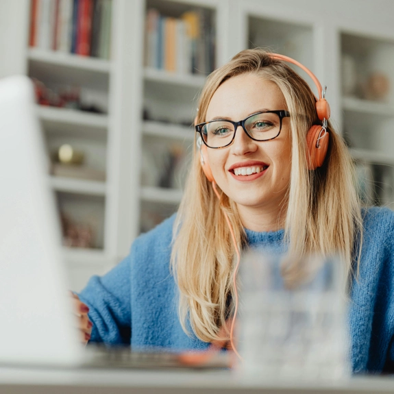 Young Blonde Woman With glasses wearing Headphones and smiling at laptop